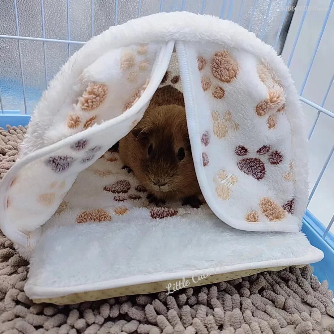 Hamaca de mascota blanca y esponjosa con huellas de conejo marrón, mostrando un pequeño conejo marrón dentro.