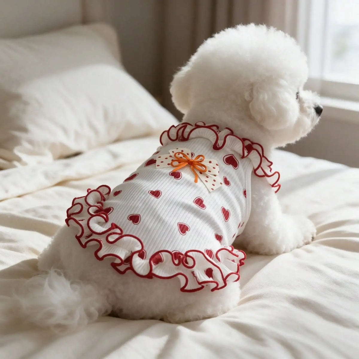A fluffy white dog wearing a red heart bow ruffle sleeveless tank top lying on white bedding.