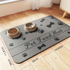 A gray pet food mat with paw and bone designs is placed on a wooden floor, showing its size relative to the room.