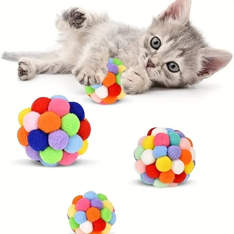 Ein kleines graues Kätzchen liegt neben einem bunten Ballset aus flauschigen Pompons.