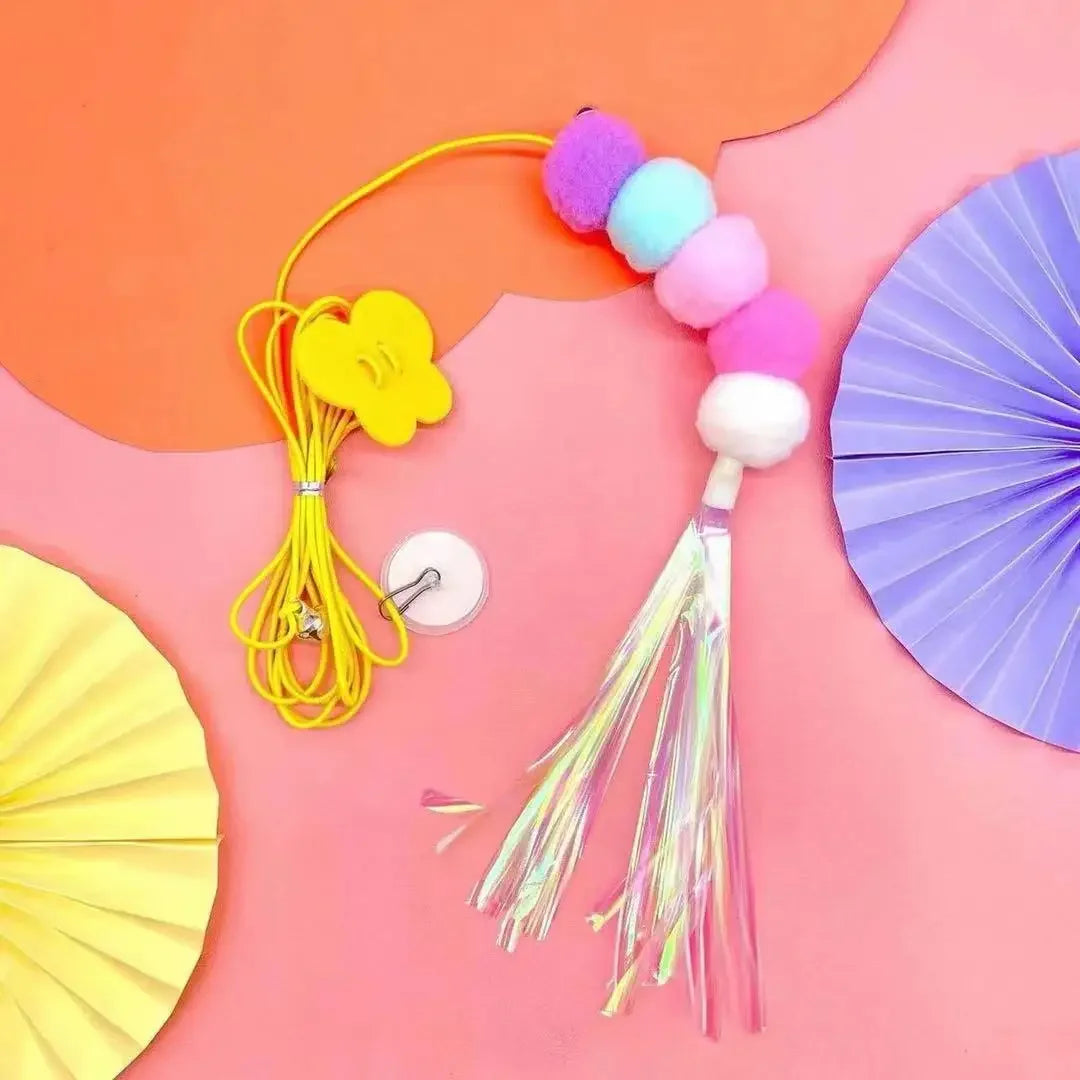 A colorful interactive hanging cat toy featuring pom-poms and a tassel, displayed on a pink background.