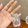 A clear, puppy-shaped pet hair keepsake keychain with a gold-colored chain and attached to a hand.