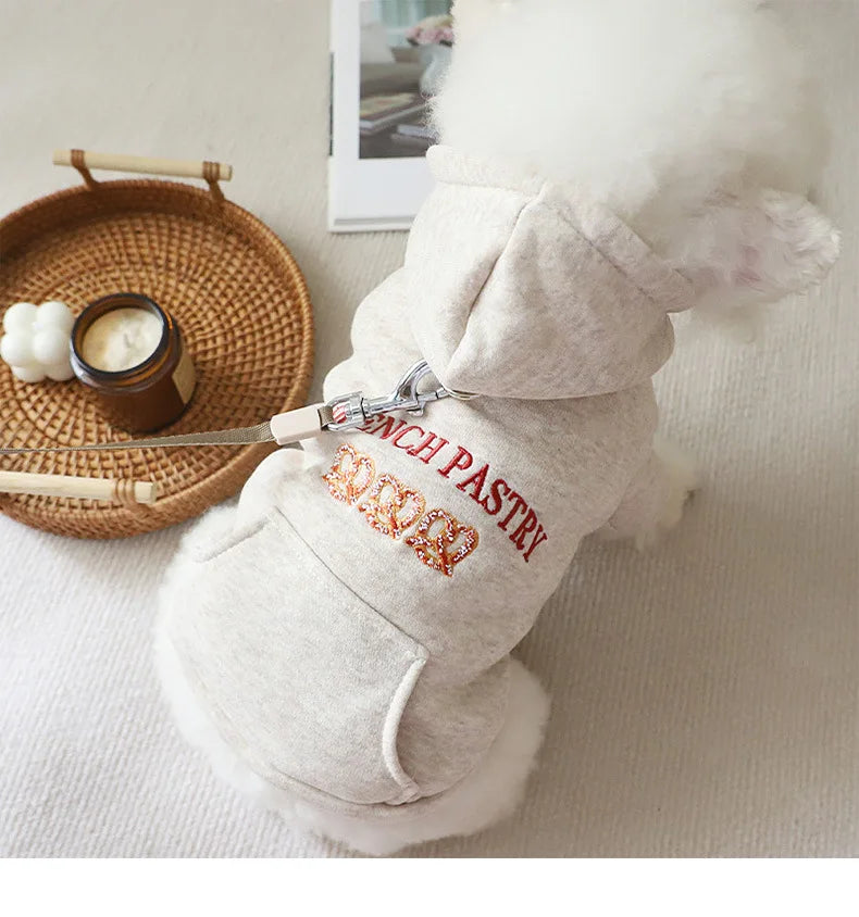 A light beige embroidered hoodie for a small dog with white fur, resting next to a wooden plate and a jar.