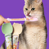 A tabby cat is being fed liquid treats using a pink squeeze spoon feeder and green silicone spoons.