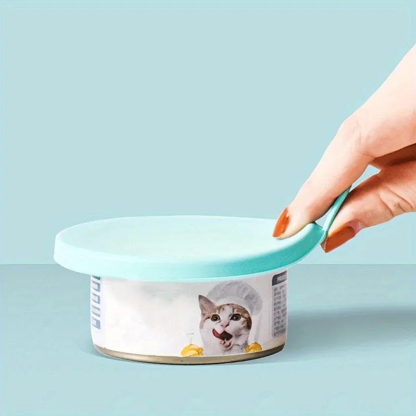 A hand is holding a light blue pet food can lid and spoon set with a cat face design.