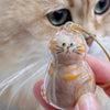 A small, clear resin charm shaped like a tabby kitten with orange stripes and green eyes, attached to a gold-colored string.