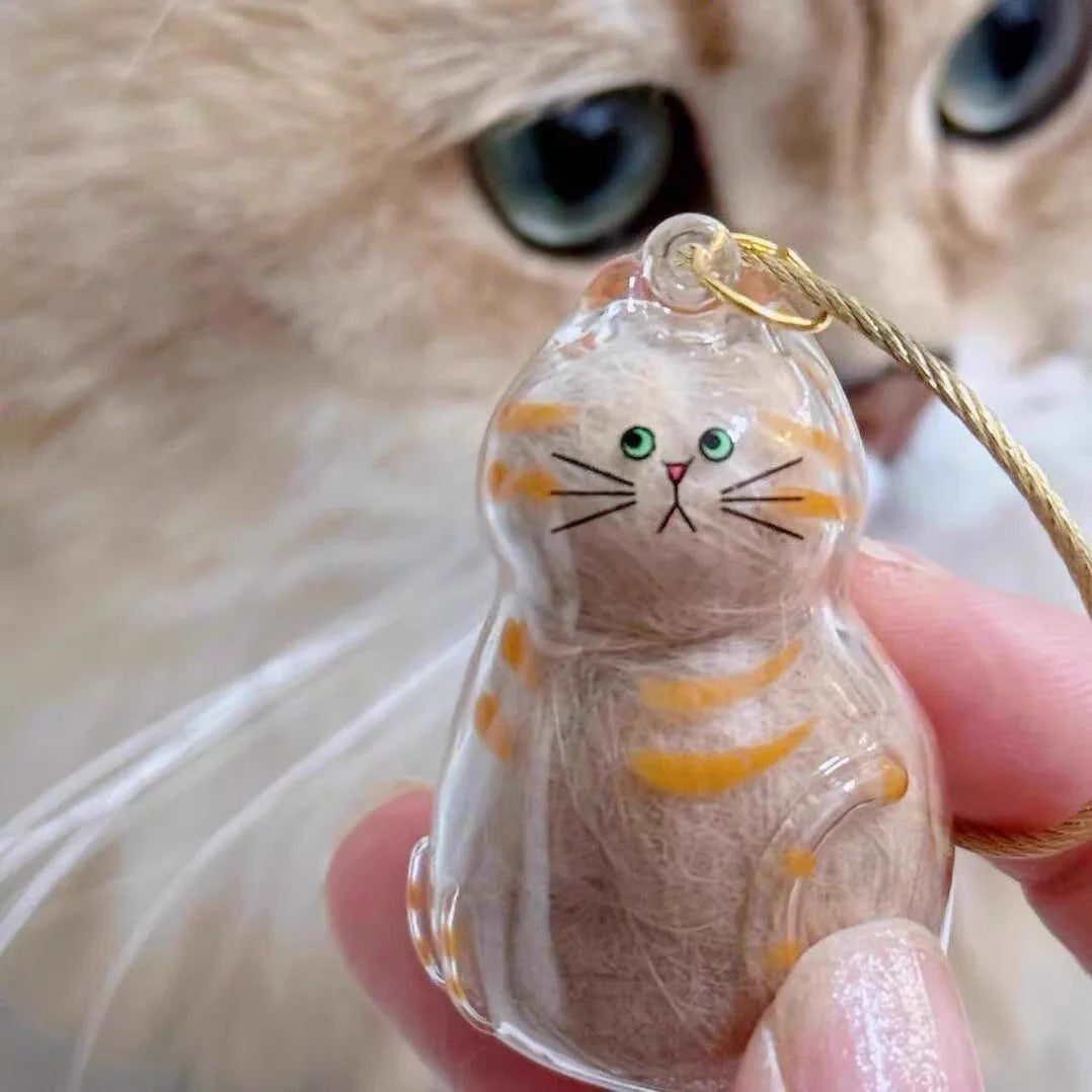 A small, clear resin charm shaped like a tabby kitten with orange stripes and green eyes, attached to a gold-colored string.
