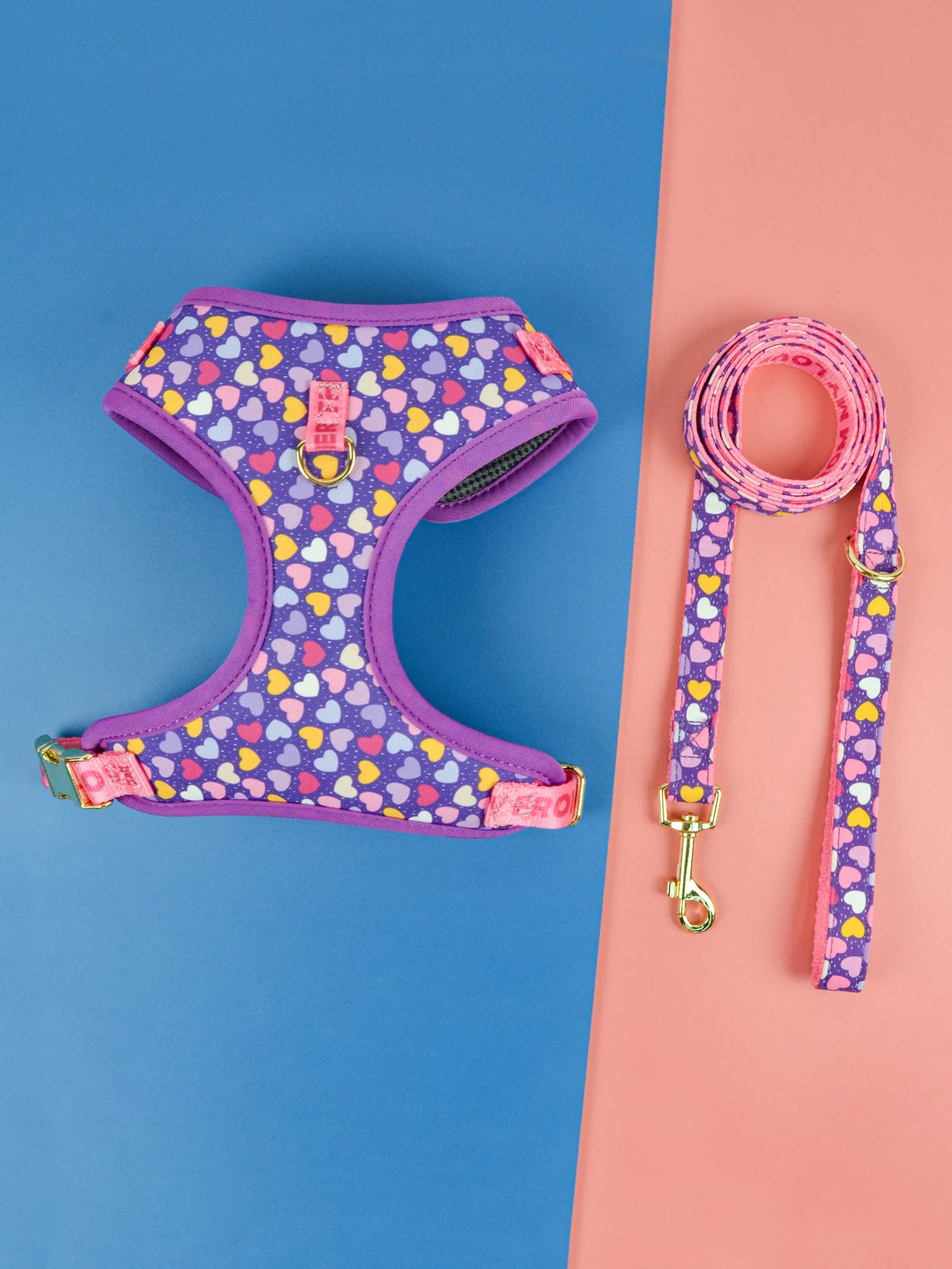 A purple pet harness with a heart pattern is displayed next to its matching pink leash on a blue and pink background.