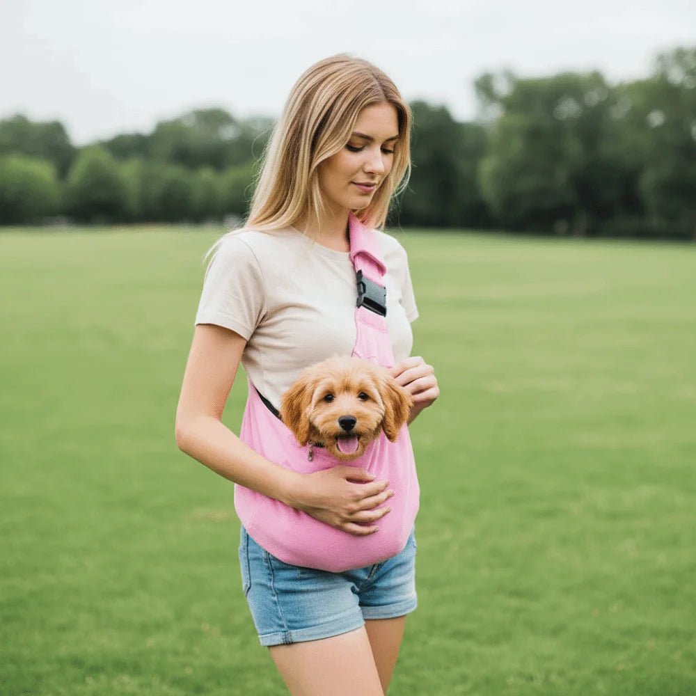 A woman wearing a pink carrier bag is holding a small golden dog outdoors in a grassy field.