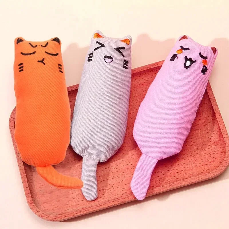 Three orange, gray, and pink cat-shaped chew pillow toys are displayed on a pink tray.