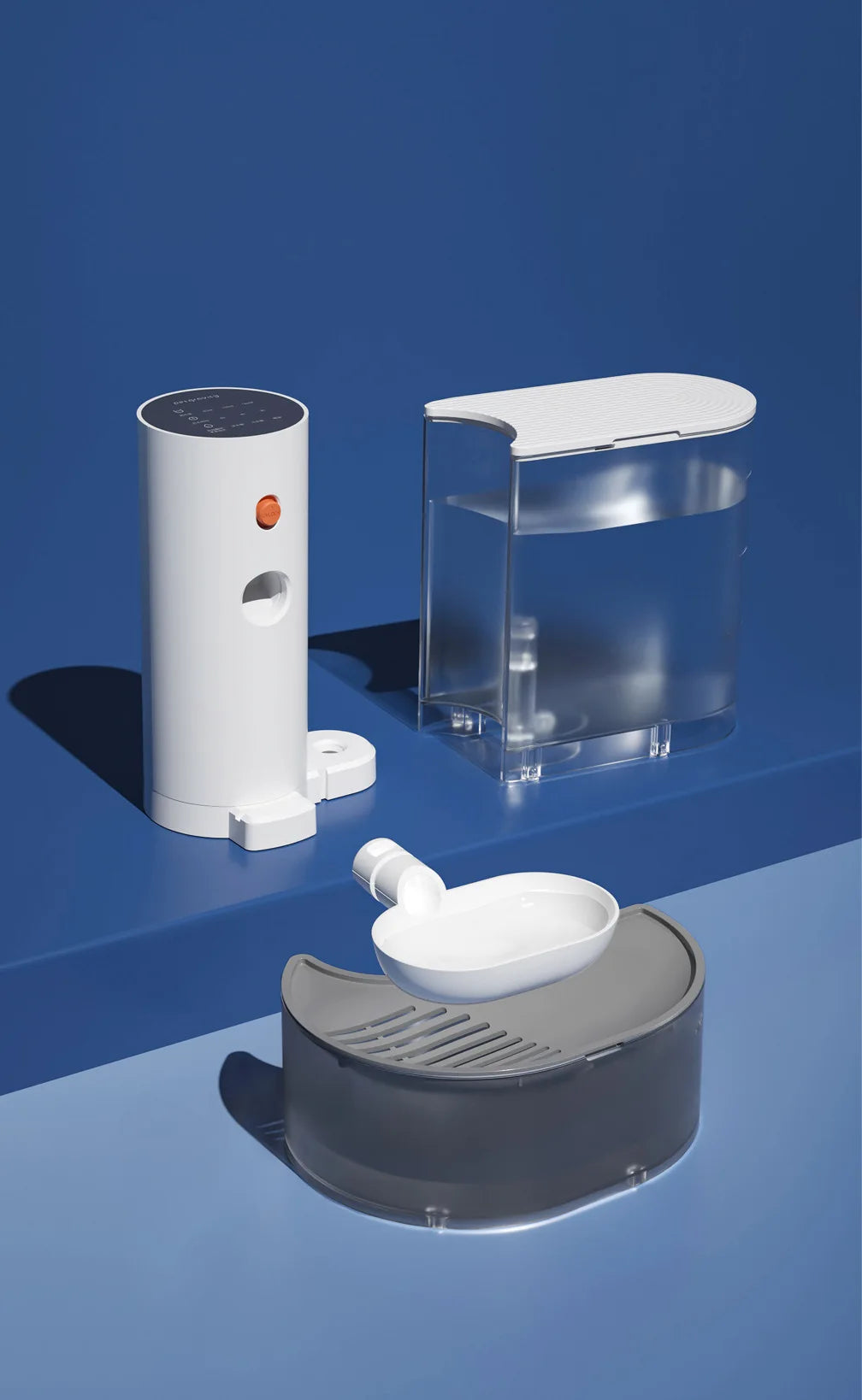 Weißer automatischer Wasserdispenser für Haustiere neben einem klaren Behälter und einer grauen Futterschale auf blauem Unter