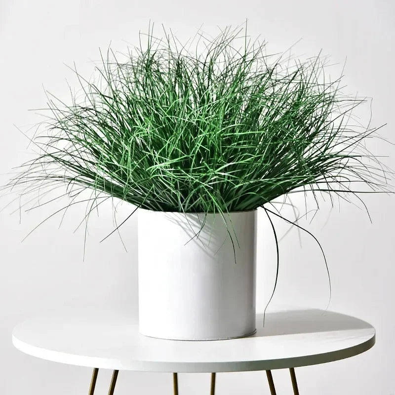 A potted faux grass bush set with green reeds and onion grass displayed on a white table.