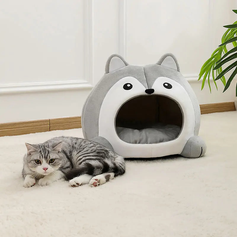 Une tanière de lit pour chat en peluche en forme de renard gris avec une entrée, à côté d'un chat tigré.