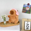 Flauschiger brauner Capybara-Plüschtier mit rosa Kapuze und Flügeln, sitzt neben einem gerahmten Bild.