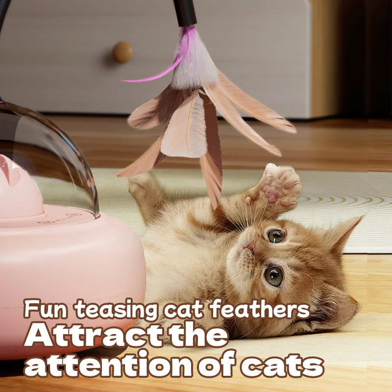 A fluffy orange kitten is lying down next to a pink automatic UFO cat teaser toy with a feather wand.