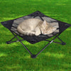 A light gray cat is relaxing in a black foldable elevated pet cot with breathable mesh on a green lawn.