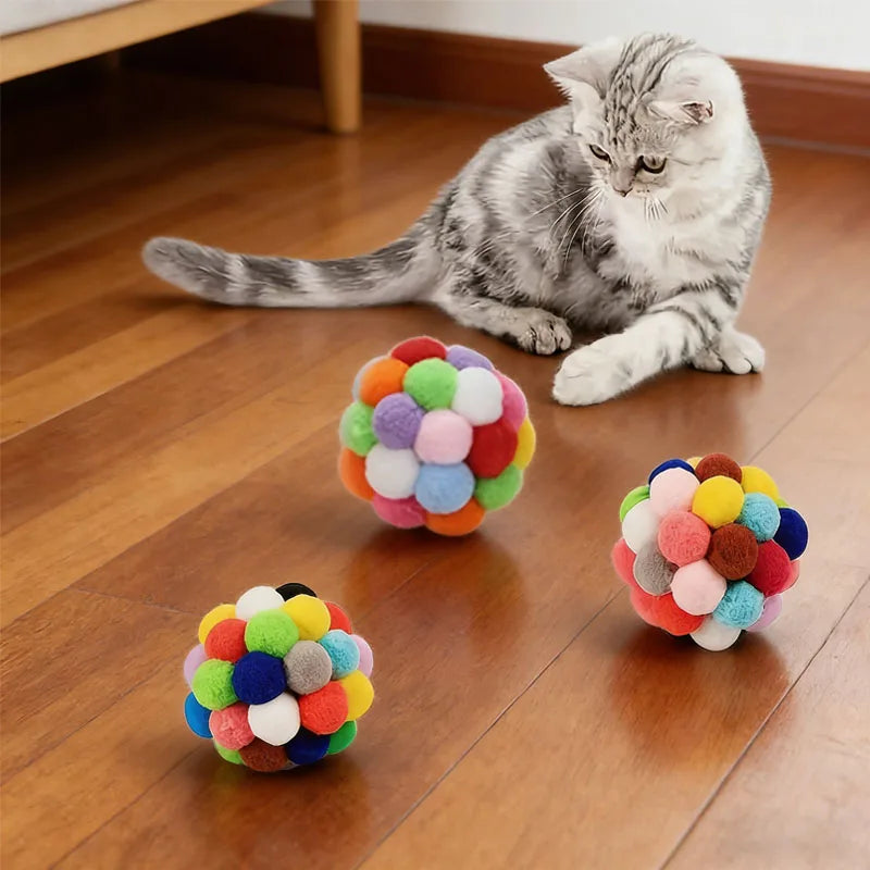 A tabby cat is sitting on a wooden floor next to several colorful, fuzzy balls used as an interactive cat toy.