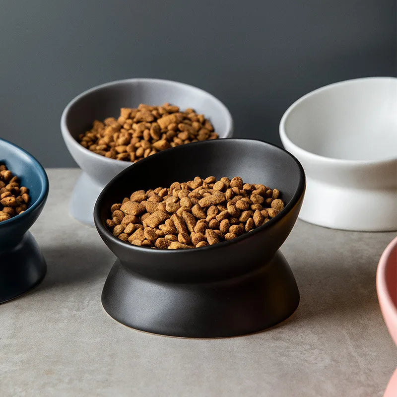 A black ceramic cat bowl with a raised stand filled with brown kibble, set against a neutral background.