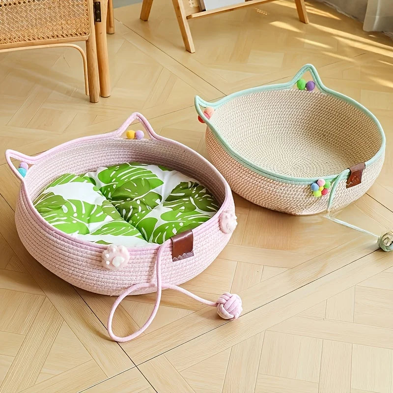 A pink hand woven rattan cat bed with a green patterned scratching surface sits on a light wooden floor.