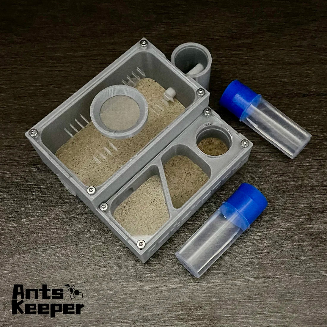 A compact plastic ant farm with a tray and two clear tubes for feeding is shown on a dark surface.