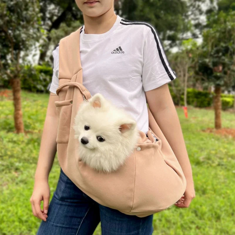 A person is wearing a beige hands-free pet sling carrier with a fluffy white dog inside, standing outdoors.