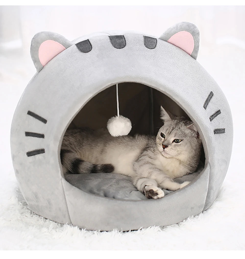 Un lit grotte en forme de chat gris avec des oreilles roses et un jouet pompon blanc à l'intérieur.