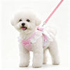 A fluffy white dog wearing a pink lace ruffle plaid harness vest with an angel heart design.
