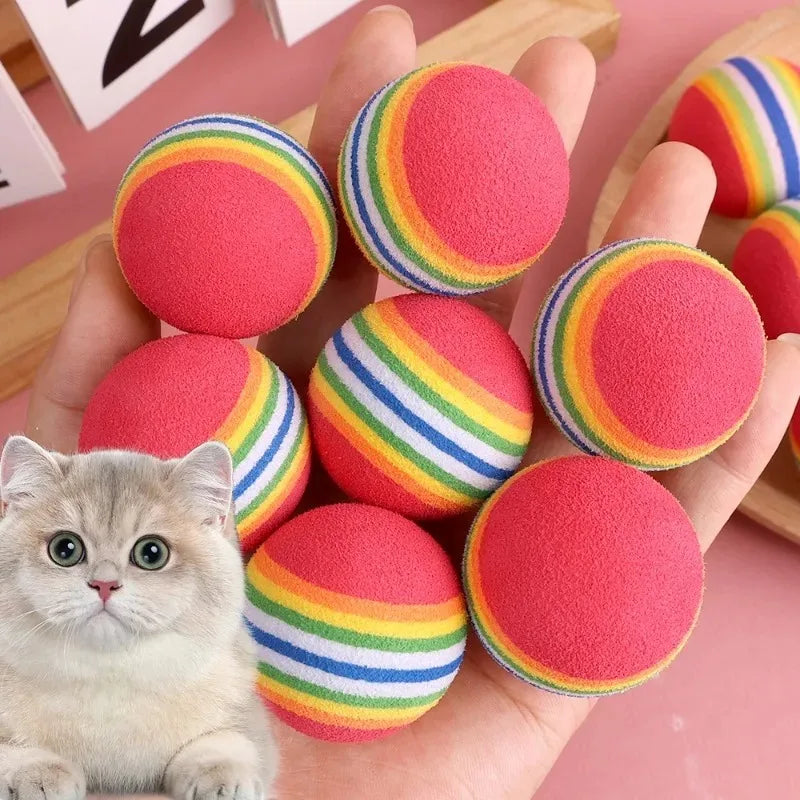 A hand holds several colorful rainbow-striped foam rattle balls, including pink and yellow, for cats and small dogs.