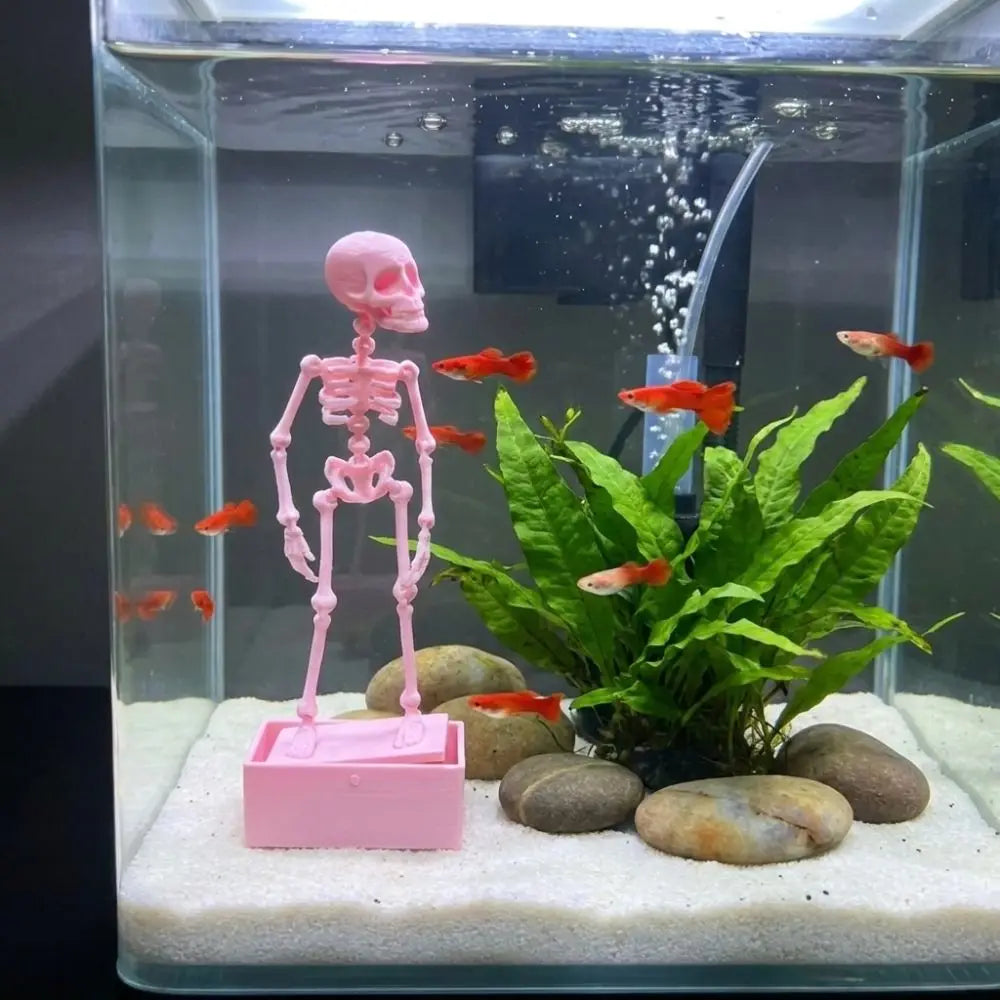 A pink skeleton figure stands on a white gravel bed surrounded by green plants and orange fish in an aquarium.