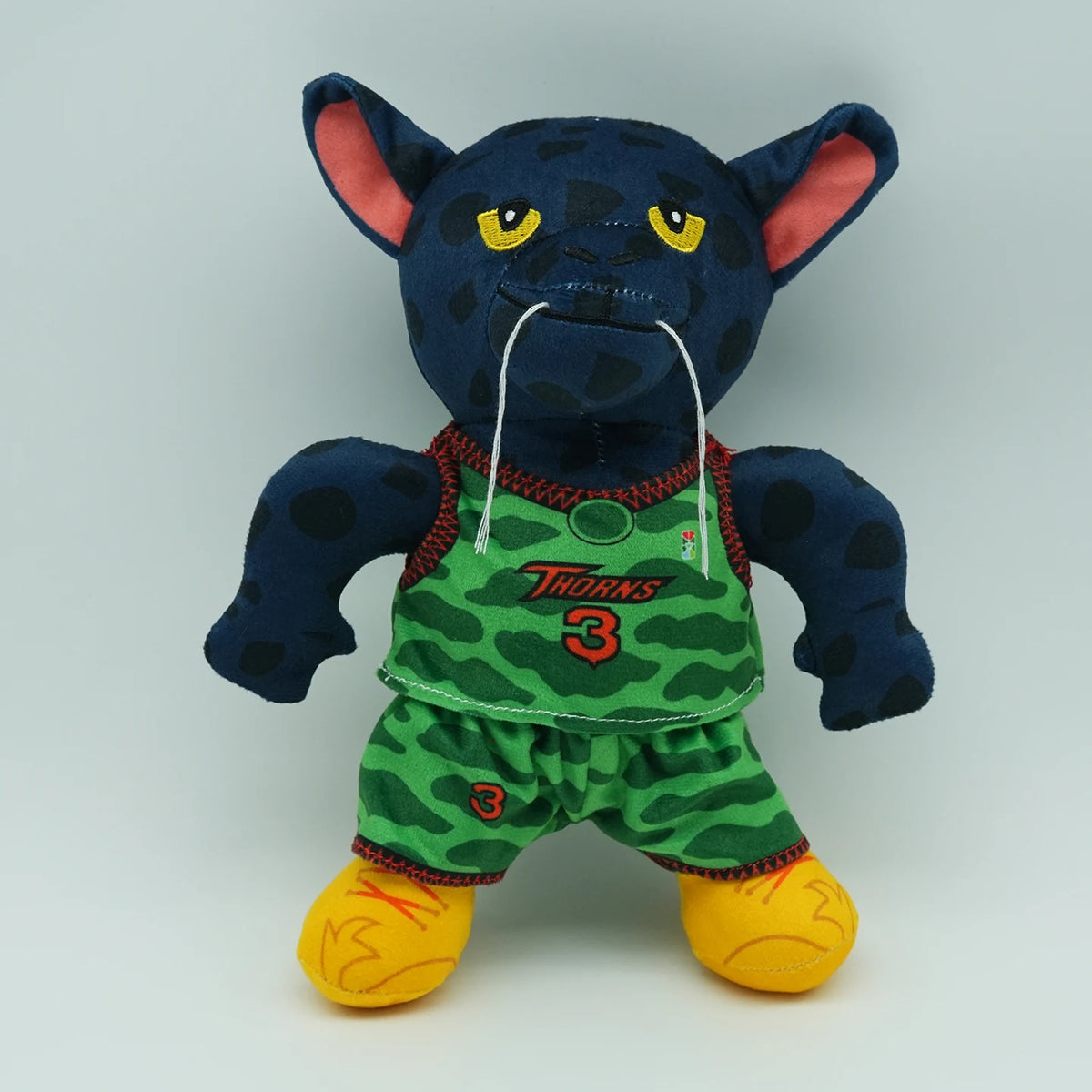 A dark blue cartoon goat plush toy with yellow eyes and green clothing.