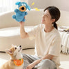 A woman is holding a blue plush dog toy while sitting on the floor with a small dog nearby.