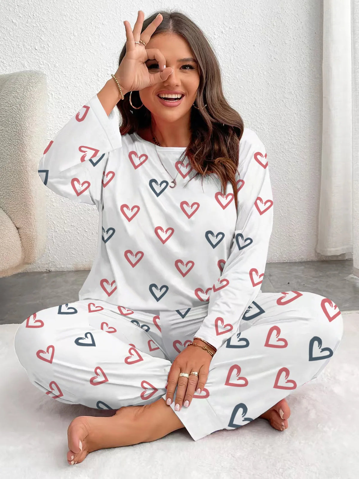A woman models a white pajama set with a repeating blue and red heart print, sitting on a white rug.