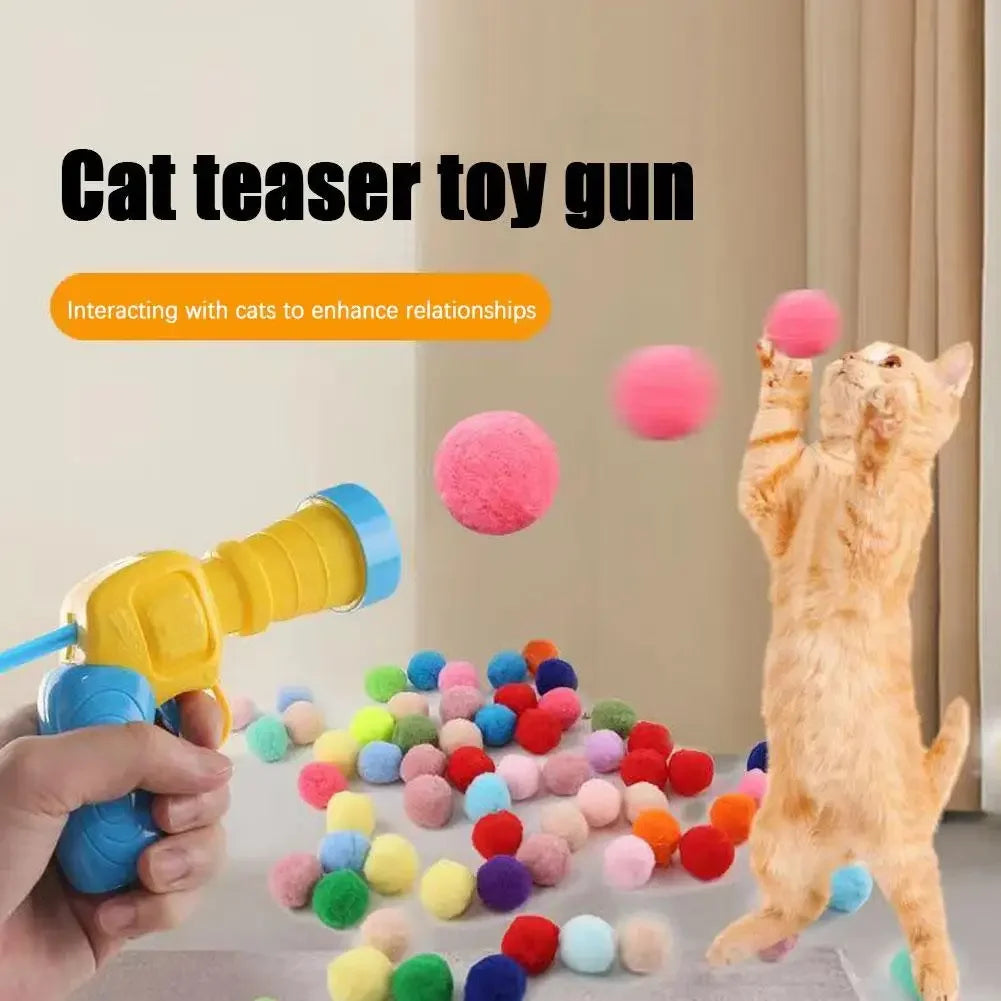 A hand is holding a yellow cat teaser toy gun, launching pink and multicolored soft plush balls toward a cat.