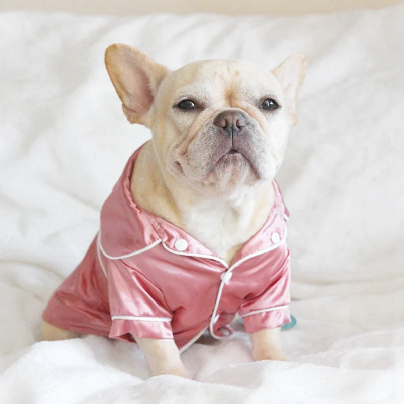 A fawn-colored dog wearing pink silk pajamas lying on white fabric.