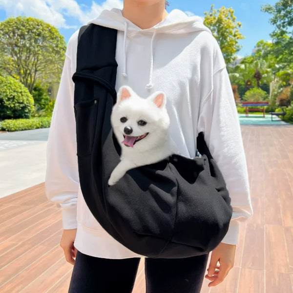 A person wearing a white hoodie is holding a black hands-free pet sling carrier with a small white dog inside.