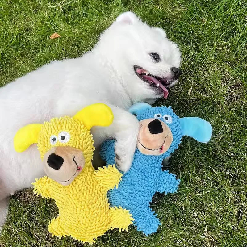 A white dog lies next to two yellow and blue plush toys on green grass.