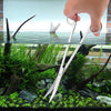 Eine Hand hält ein Set zum Reinigen eines Aquariums aus Edelstahl über einem Glasbehälter mit Wasserpflanzen und Abfällen.