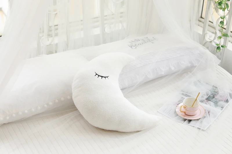 A white crescent moon shaped plush pillow with a delicate face is resting on a white bed with sheer white fabric.