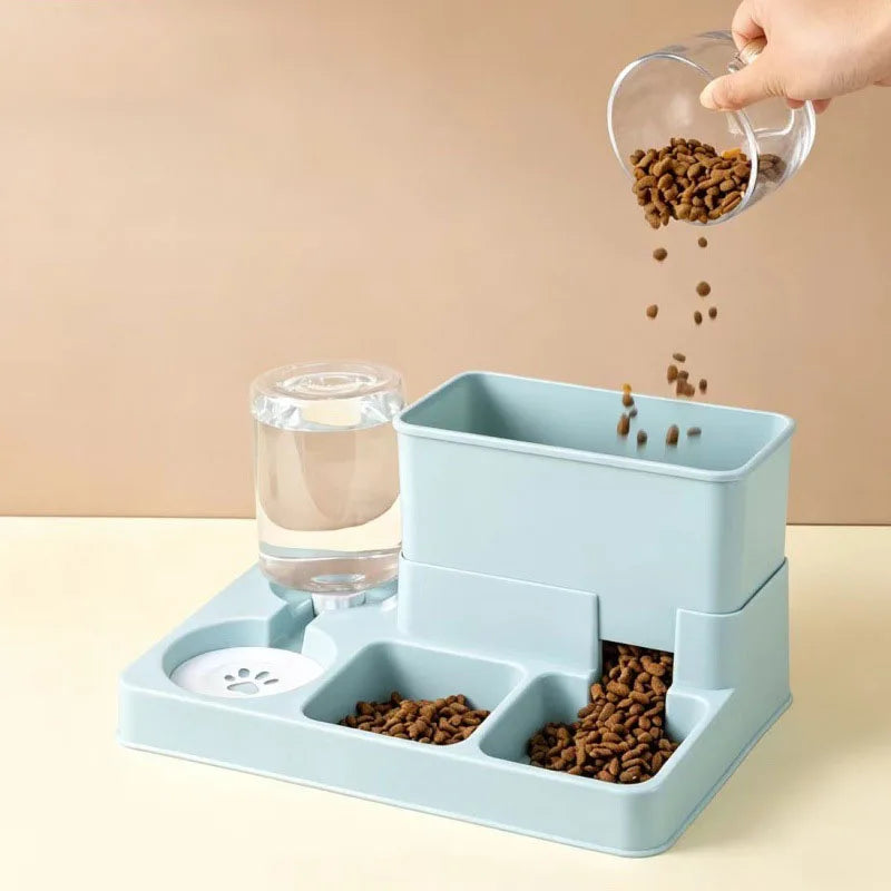 A hand is pouring dry cat food into a blue plastic feeder, next to a glass water fountain.