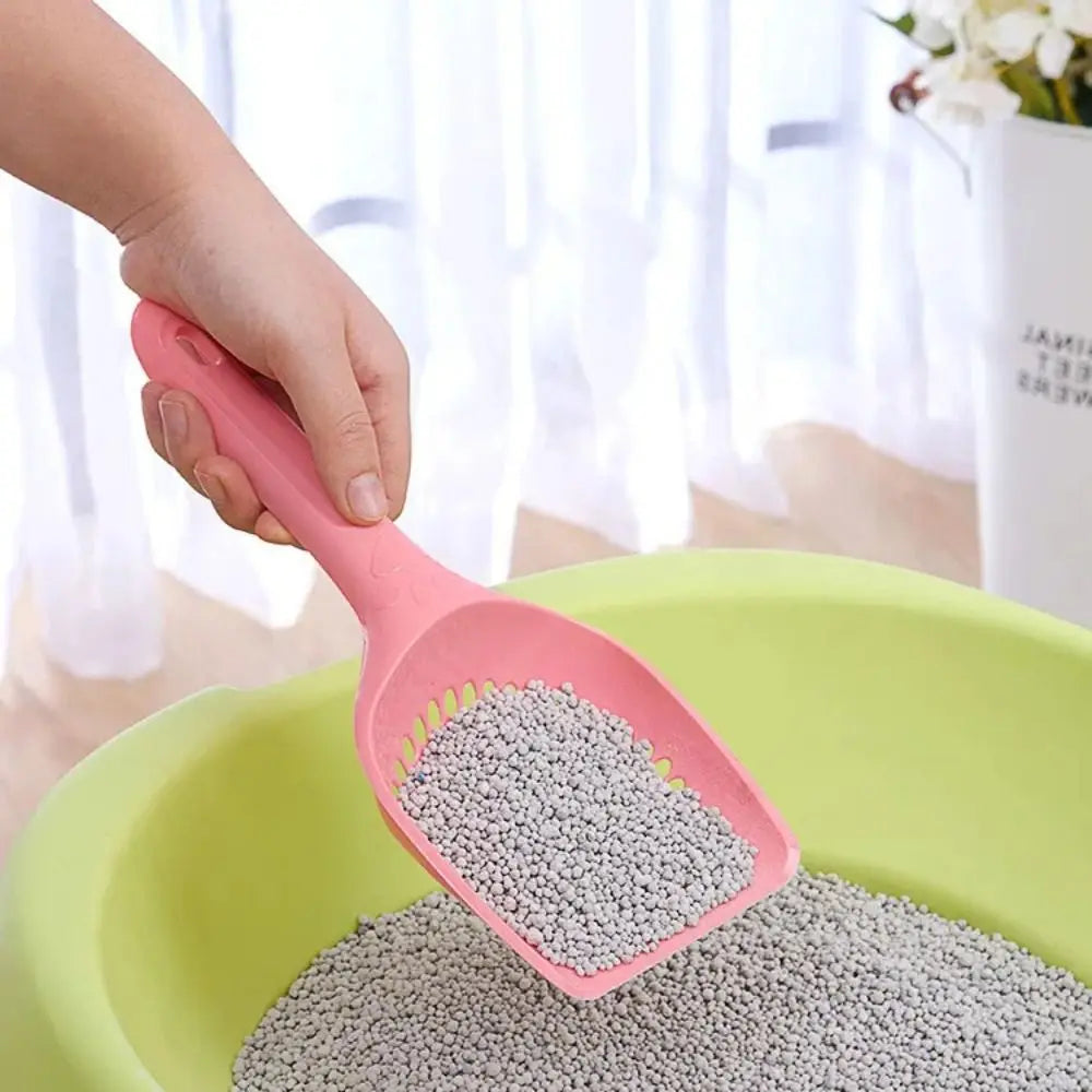 A hand is using a pink durable cat litter scoop with sifting slots to transfer gray litter into a green bowl.