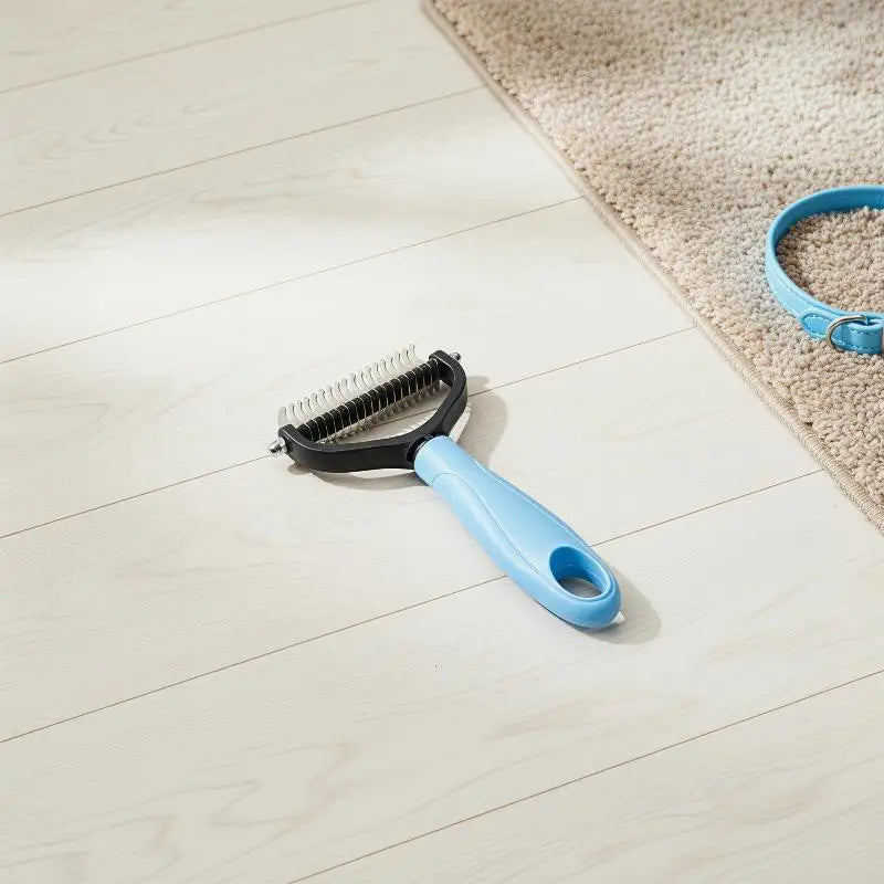 A black and light blue 2-in-1 pet dematting and deshedding brush lying on a light wooden floor next to a beige rug.