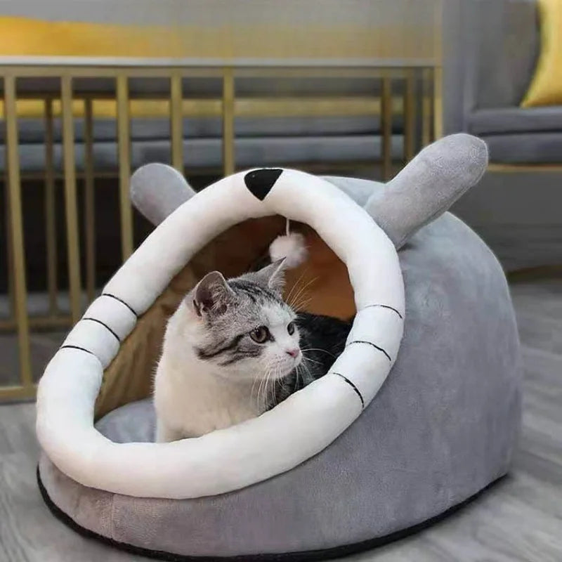 A domestic cat peeks out of a round, soft gray pet cave bed with a white, rolled-up entrance.