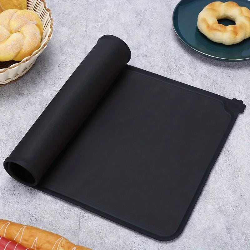 Black silicone pet feeding mat with raised edges placed on a gray surface next to baked goods.