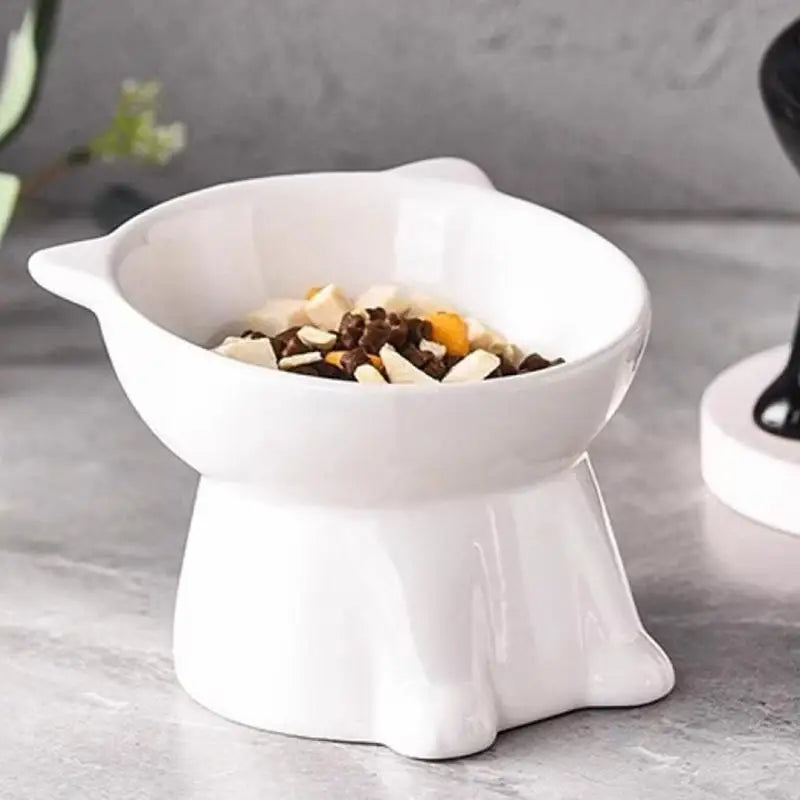A white elevated slanted cat bowl with an anti-tip base filled with dry food is shown on a gray surface.