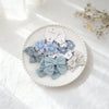 Collection de nœuds pour cheveux et accessoires en tissu bleu clair et gris présentés sur une assiette blanche.