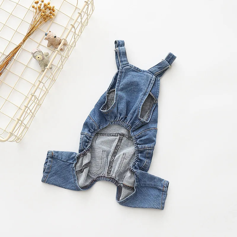 A denim dog jumpsuit costume for small to medium breeds lying on a white surface next to a wire basket.
