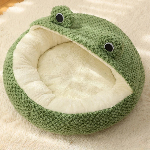 Nido de mascota de peluche verde con forma de rana y interior blanco, sobre una superficie de pelaje claro.