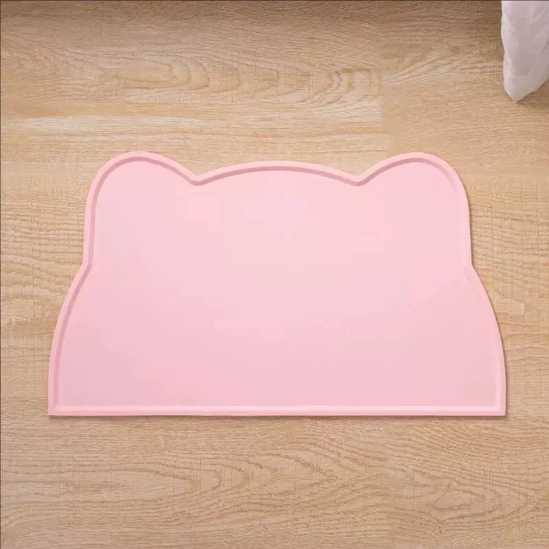 A pink silicone cat face pet feeding mat for food and water bowls placed on a wooden surface.