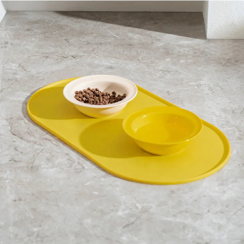 A yellow non-slip feeding mat with two ceramic bowls for pet food is placed on a gray countertop.