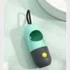 A light teal capsule dog waste bag dispenser with a black handle and a yellow button, resting on a white surface.